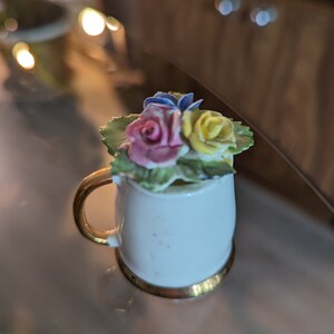 Peut inclure: Une tasse en céramique blanche avec un bord et une poignée dorés, décorée d'un bouquet de fleurs en porcelaine rose, jaune et bleue.
