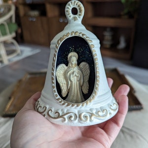 May include: White ceramic bell with a gold trim and an angel figurine inside. The angel is wearing a gold gown and has its hands clasped in prayer. The bell has a blue background with gold glitter.