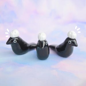 May include: Three glossy black seal figurines, each wearing a small white hat. The seals have white eyes and are arranged on a pastel-coloured background. The figurines are whimsical and decorative.
