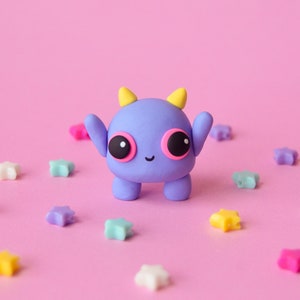 May include: A purple monster with yellow horns and pink eyes, surrounded by colourful star-shaped decorations on a pink background.