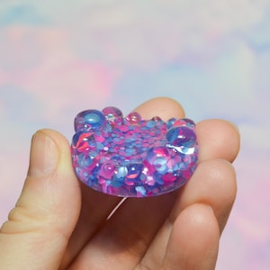 May include: A small, colorful resin object with a rounded shape. The object is filled with pink, blue, and purple glitter and small, clear, bubble-like embellishments. The object is held in a hand against a soft, pastel background.