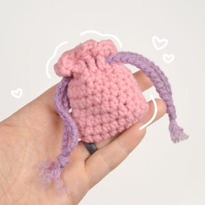 May include: A small, handmade crochet pouch in soft pink with a drawstring closure. The drawstring is a gradient of purple hues. The pouch is held in a hand against a white background, with white heart doodles.