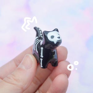May include: A small, black cat figurine with a skeleton design. The cat has a white skull-shaped face and white bones painted on its body. The figurine is held in a hand against a pastel background.