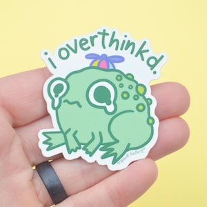 May include: A die-cut sticker showing a cartoon frog with the words "i overthinkd." The frog is light green with a small hat and is crying. The sticker is held in a hand against a yellow background.