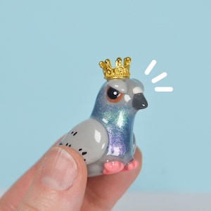 May include: A miniature pigeon figurine with a gold crown, featuring iridescent blue and grey feathers, pink feet, and a grumpy expression. The figurine is held by a hand against a light blue background.