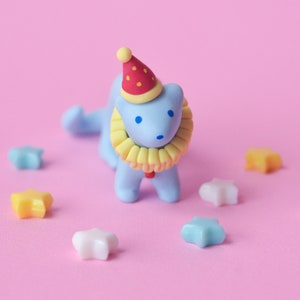 Peut inclure: Une figurine en argile bleue d'un lion portant un chapeau rouge et blanc et un collier jaune. Le lion est entouré de petites décorations en forme d'étoile colorées sur un fond rose.