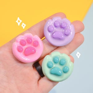 May include: Three colourful paw-shaped resin charms in pink, purple, and green. Each charm features a paw print design with rounded pads and claws. The charms are held in a hand against a yellow and blue background.