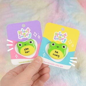 Könnte beinhalten: Zwei handgefertigte Tonpins mit einem grünen Froschdesign. Ein Pin hat "she her" auf einem gelben Oval, der andere "he they". Beide Pins befinden sich auf lila und gelben Karten mit dem Text "sploot factory".