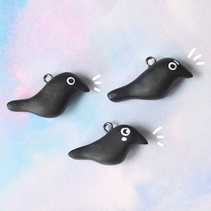 May include: Three black bird-shaped charms with white accents and silver jump rings. Each charm has a white eye and a stylized white speech bubble. The charms are on a pastel blue and purple background.