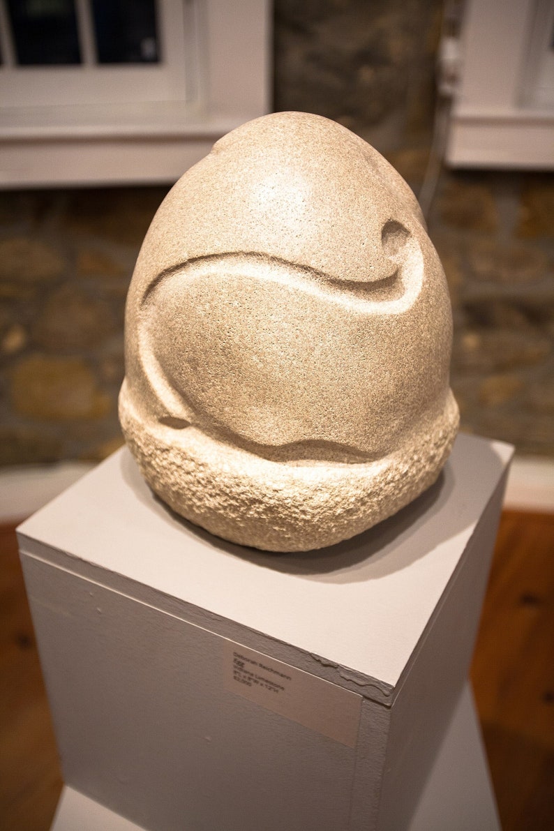 May include: A white stone sculpture of an egg with a carved design. The sculpture is displayed on a white pedestal.