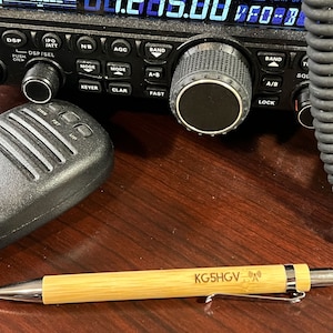 May include: A wooden pen with a silver clip and a black and silver tip. The pen has the text "KG5HGV" engraved on the side. The pen is lying on a wooden surface.