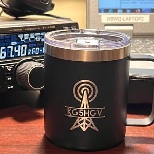 Puede incluir: Taza aislada negra con borde y asa plateados. La taza presenta un gráfico plateado de una torre de radio con el indicativo KG5HGV. Un transceptor de radio y un portátil están en el fondo.