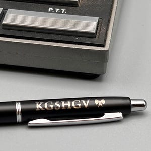Puede incluir: Primer plano de un bolígrafo negro con el texto "KGSHGV" en letras doradas. El bolígrafo tiene un clip y una punta plateados. La imagen también muestra parte de un dispositivo gris con las letras "P.T.T.".