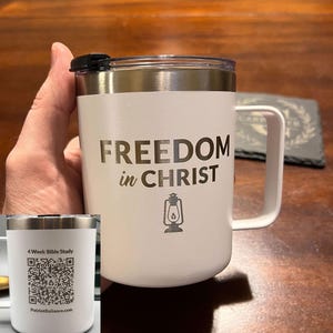 May include: White travel mug with a handle and a black lid. The mug features the text "FREEDOM in CHRIST" and a lantern graphic. A smaller image shows a QR code and the text "4 Week Bible Study" and "PatriotReliance.com".