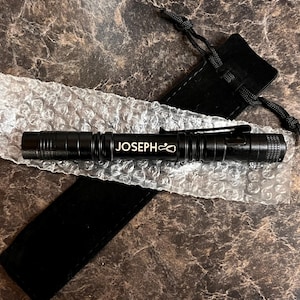 May include: A black flashlight with a silver inscription that reads "JOSEPH" and a small symbol. The flashlight is lying on a white bubble wrap surface and a black pouch.