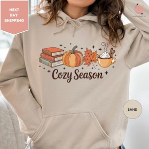 May include: Sand-coloured hoodie with a fall-themed graphic. The design features a stack of books, a pumpkin, a maple leaf, and a mug of hot cocoa with cinnamon sticks. The words "Cozy Season" are printed below the graphic.