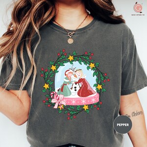 A woman wearing a gray t-shirt with a Christmas-themed graphic design featuring two people, a snowman, and a wreath.