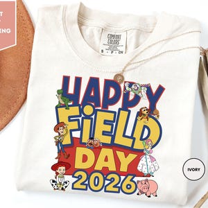 A white t-shirt with a colorful graphic design featuring characters from the Toy Story movie, along with the text "Happy Field Day 2026".