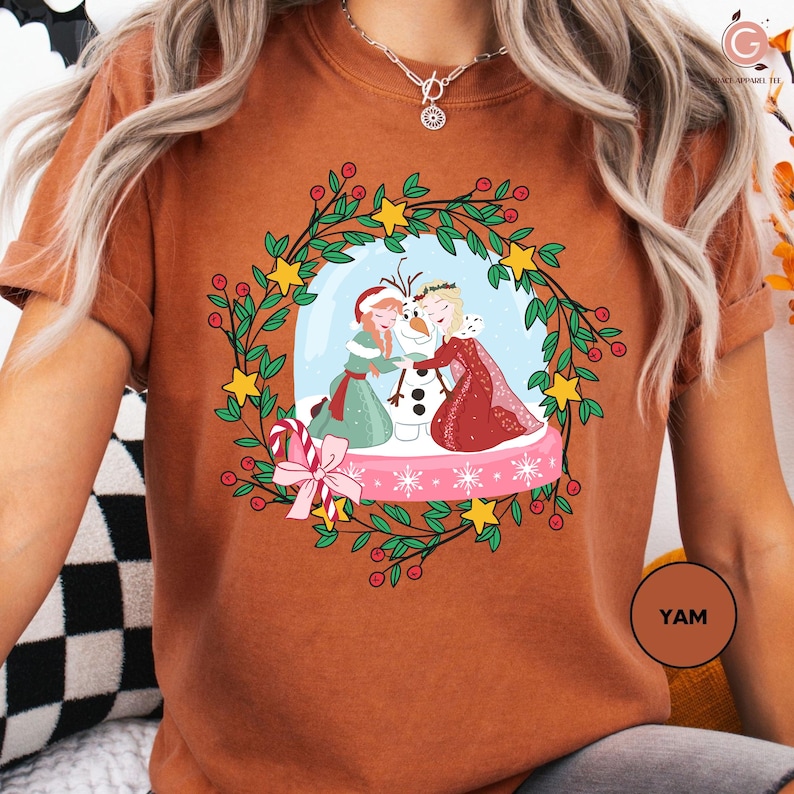 A woman wearing a brown t-shirt with a Christmas-themed graphic design featuring two women, a snowman, and a wreath.