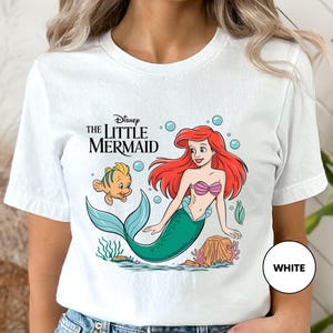 a woman wearing a white shirt with the little mermaid on it