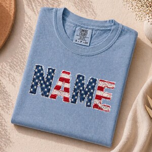 Puede incluir: Una camiseta azul claro con cuello redondo y etiqueta de la marca Comfort Colors. En la parte delantera aparece la palabra NAME en letras grandes de estilo desgastado, con un patrón de estrellas blancas sobre fondo azul y rayas rojas.