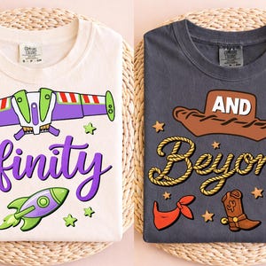 Op de afbeelding: Twee opgevouwen t-shirts met cartoonafbeeldingen. Eén is crèmekleurig met een paars "infinity"-ontwerp en een raket. De andere is donkergrijs met een cowboyhoed en de woorden "And Beyond".