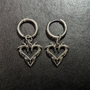 May include: A pair of silver hoop earrings with a heart-shaped charm. The heart charm has a detailed, tribal-inspired design.