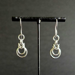 Może przedstawiać: Para srebrnych kolczyków o unikalnym designie. Każdy kolczyk posiada mały, dekoracyjny element nad dwoma połączonymi okrągłymi obręczami. Kolczyki są prezentowane na czarnym stojaku na ciemnym tle.