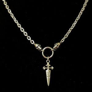 May include: Silver necklace with a dagger pendant. The chain is linked to two skull charms. The dagger pendant has an intricate design. The necklace is set against a black backdrop.