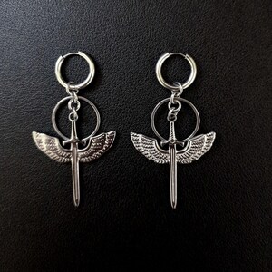 May include: A pair of silver-colored earrings. Each earring features a sword design with detailed wings, suspended from a hoop and a smaller circular element. The earrings are displayed against a dark background.