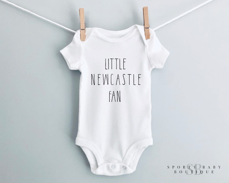 May include: White baby bodysuit with the words "LITTLE NEWCASTLE FAN" printed in black. The garment is suspended from a washing line with wooden pegs. The backdrop is a pale grey.