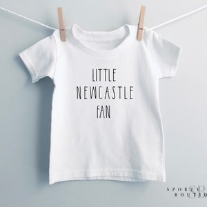 May include: White t-shirt with the text "LITTLE NEWCASTLE FAN" in black, suspended from a clothesline with a wooden peg. The shirt has a crew neck. The words "SPORTY BABY BOUTIQUE" are at the bottom right.