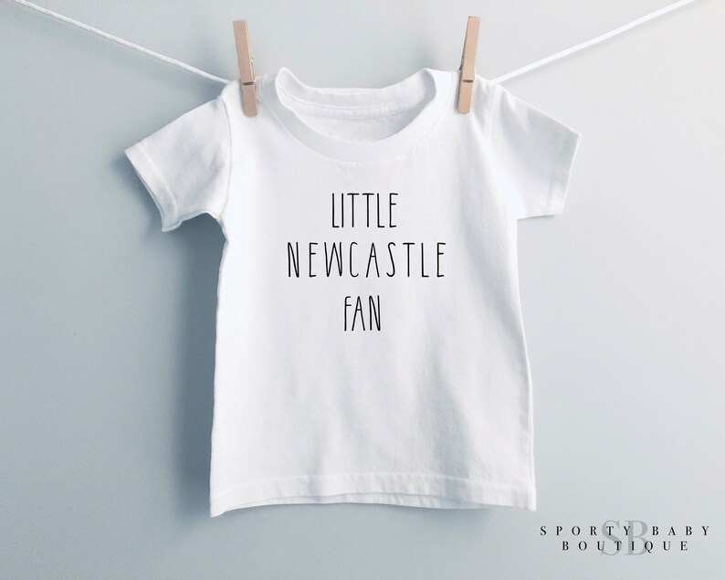 May include: White t-shirt with the text "LITTLE NEWCASTLE FAN" in black, suspended from a clothesline with a wooden peg. The shirt has a crew neck. The words "SPORTY BABY BOUTIQUE" are at the bottom right.