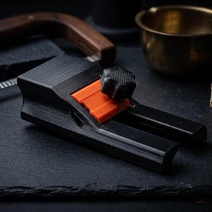 May include: A black soap cutter with an orange adjustable guide and a black knob, positioned near a wooden soap slicer, a small brass bowl, and two stacked bars of soap with dried flower petals scattered on the surface.