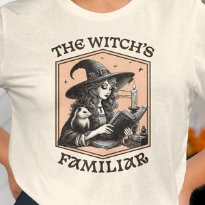 Może przedstawiać: Biały T-shirt z czarno-białym obrazkiem przedstawiającym czarownicę czytającą książkę z małym zwierzęciem u boku. Na koszulce nadrukowany jest tekst "The Witch's Familiar".