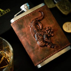 Könnte beinhalten: Eine braune Lederflasche mit einem silbernen Metalldeckel und einem detailliert geprägten Tigerdesign. Die Flasche befindet sich auf einer dunklen Oberfläche.