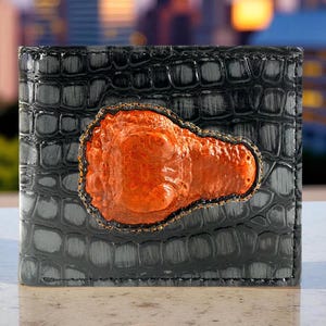Könnte beinhalten: Schwarze Ledergeldbörse mit strukturiertem Krokodilmuster. Die Geldbörse hat ein orangefarbenes, strukturiertes Design in der Mitte, das von einer goldfarbenen Umrandung eingefasst ist. Die Geldbörse ist rechteckig und scheint geschlossen zu sein.