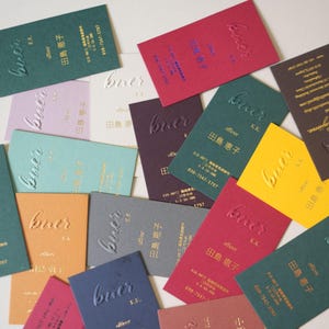 May include: A collection of business cards in various colors, including green, red, and yellow. Each card features the embossed word "buer" and Japanese text. The cards are rectangular and arranged in a scattered pattern.