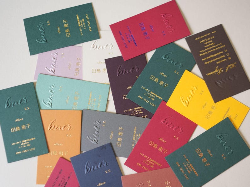 May include: A collection of business cards in various colors, including green, red, and yellow. Each card features the embossed word "buer" and Japanese text. The cards are rectangular and arranged in a scattered pattern.