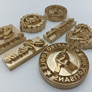 May include: A set of nine gold-colored metal stamps with various designs, including a heart, a swirl, a hand, and the words "Crafted" and "Artisan".
