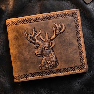 Può includere: Un portafoglio pieghevole in pelle marrone con un design a testa di cervo in rilievo al centro. Il portafoglio è incorniciato da un motivo decorativo a treccia lungo i bordi, che mostra una finitura invecchiata con texture e sfumature visibili.