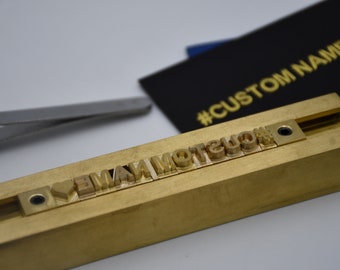Op de afbeelding: Een messing stempelblok met de woorden "I LOVE MY NAME" in reliëfletters. Het blok wordt gebruikt om aangepaste reliëfdesigns op leer of andere materialen te maken. Het blok ligt op een wit oppervlak.