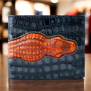 Könnte beinhalten: Ein zweifach gefaltetes Portemonnaie mit dunkelblauem, strukturiertem Äußeren und einem orangefarbenen Alligatordesign in der Mitte. Das Alligatordesign ist mit einem Kontrastfaden auf das Portemonnaie genäht. Das Portemonnaie ist aus Leder.
