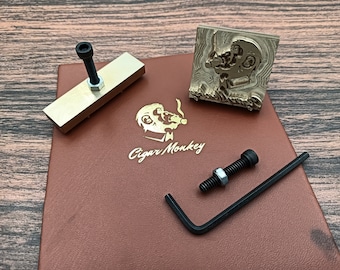 Op de afbeelding: Een messing stempel met een ontwerp van een cartoon-aap die een sigaar rookt. De stempel bevindt zich op een bruin leren oppervlak met de woorden "Cigar Monkey" in goud gedrukt. Er is ook een messing stempel met een handvat en een moersleutel.