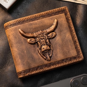 May include: A square brown leather wallet featuring a prominent embossed bull head design in the centre. The wallet is framed by a decorative Greek key border pattern stitched along the edges. The leather has a distressed, weathered finish.