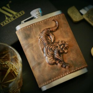 May include: A brown leather flask with a stitched edge and a silver metal body. The flask features an embossed design of a tiger.