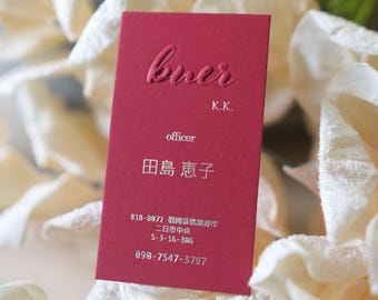 Puede incluir: Una tarjeta de visita burdeos con texto en relieve. La tarjeta dice "buer" en la parte superior, seguido de "K.K." y "officer". Debajo, se ven caracteres japoneses y un número de teléfono. La tarjeta está sobre un fondo suave y borroso.