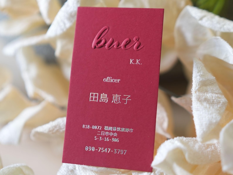 May include: A burgundy business card with embossed text. The card reads "buer" at the top, followed by "K.K." and "officer." Below, Japanese characters and a phone number are visible. The card is set against a soft, blurred background.