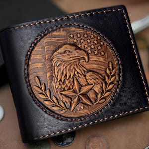 May include: Black leather wallet with a detailed, embossed design. The design features an eagle, a star, and the American flag. The wallet has a stitched border and a bi-fold design. The overall theme is patriotic.