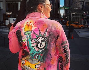 Könnte beinhalten: Eine rosafarbene Jeansjacke mit handbemaltem Design, das die Freiheitsstatue, ein gelbes Taxi und die Worte "From Brooklyn" auf dem Ärmel zeigt.